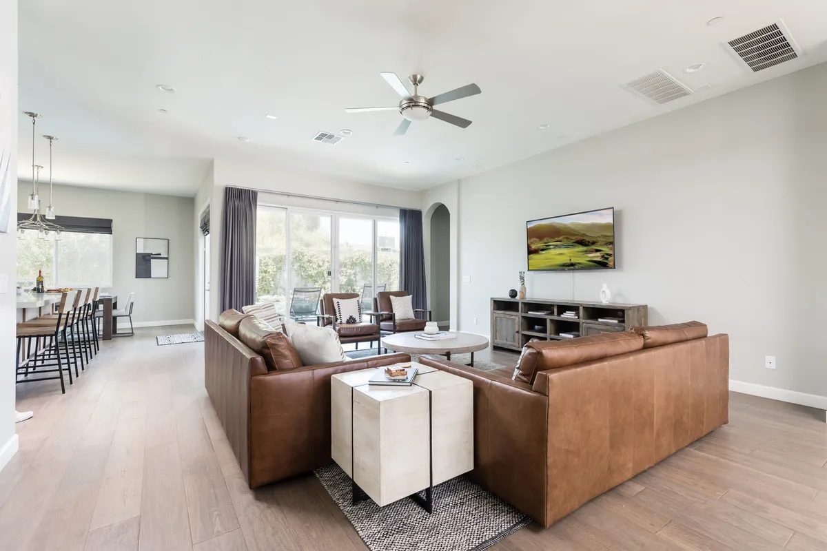 A spacious modern living room with plush leather sofas arranged around a sleek coffee table, facing a large wall-mounted TV. Expansive windows fill the space with natural light and offer a view of the outdoors, enhancing the inviting and communal atmosphere.
