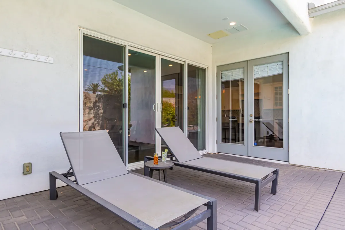 An elegant patio scene with comfortable lounge chairs, a table set with drinks, and large glass doors connecting the patio to the home’s interior. The mood is calm and inviting, ideal for sunbathing, reading, or socializing.