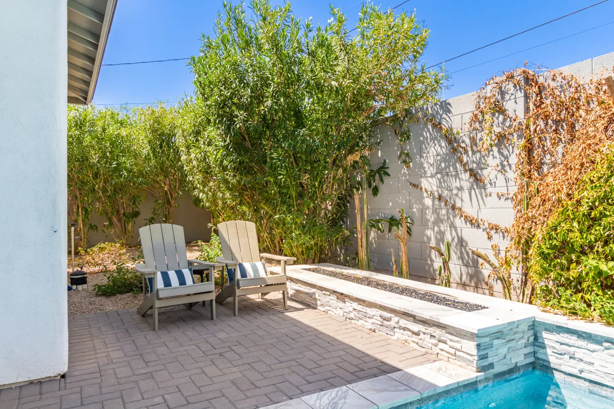 The image showcases a serene outdoor area with Adirondack chairs, a stone bench, and a sparkling swimming pool set amidst lush greenery. The arrangement suggests ample space for relaxing, entertaining, or gathering with family and friends.