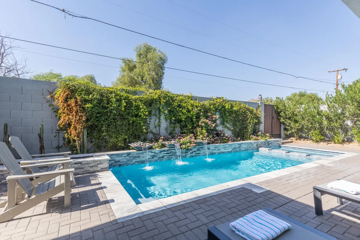 A serene backyard scene featuring a sparkling swimming pool with built-in water features. Surrounding the pool are plush outdoor seating areas and lounge chairs, all enveloped by lush greenery. The inviting ambiance suggests both daytime relaxation and evening gatherings by the pool.
