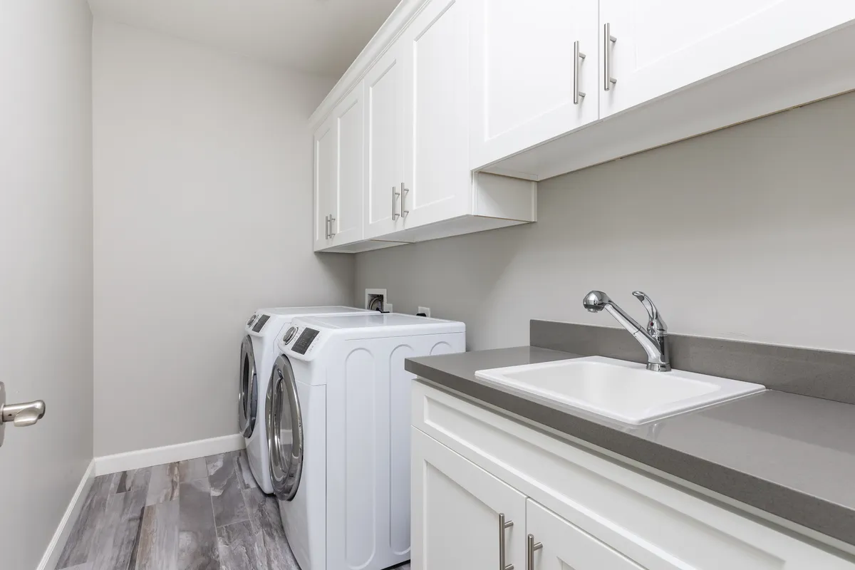 A bright, organized laundry room featuring a front-load washing machine and dryer, a utility sink, and upper cabinets for storage. The tidy layout and clean surfaces highlight a sense of order and thoughtful preparation.
