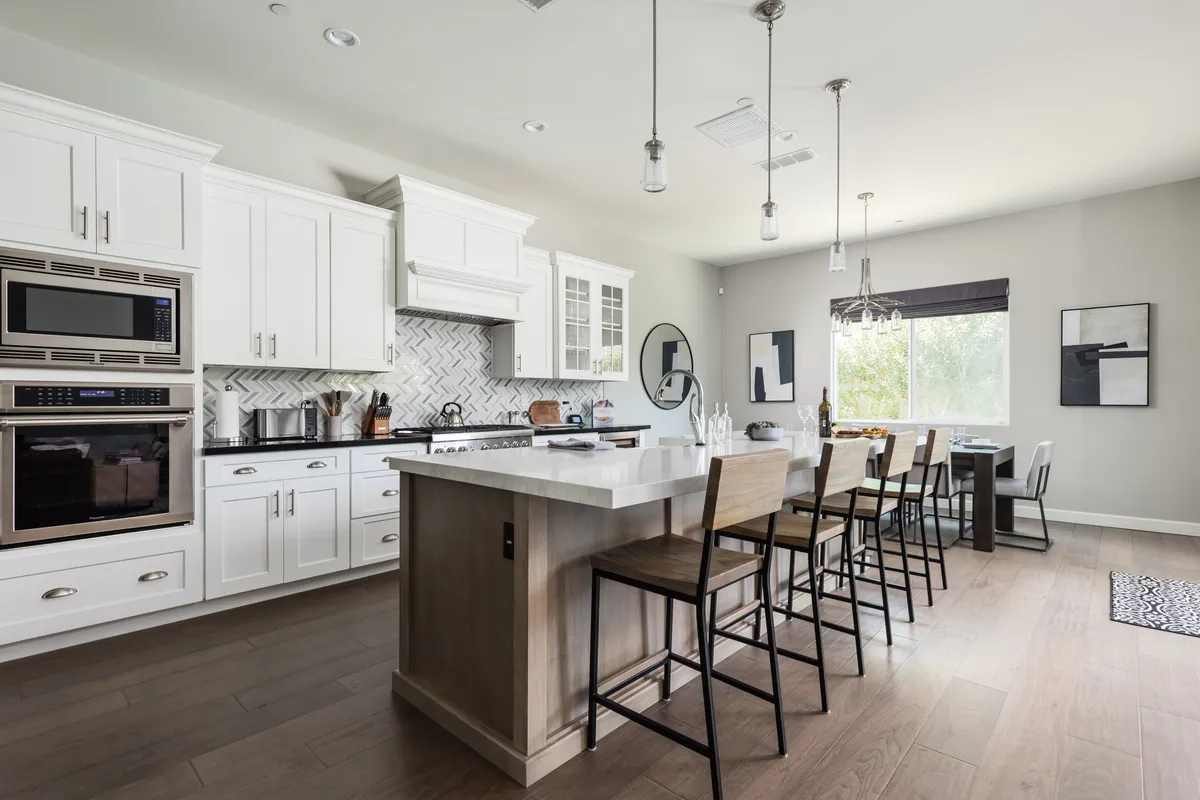 A spacious, sunlit kitchen with a large island, stainless steel appliances, a chevron tile backsplash, and a dining table by the window. The area is designed for both meal preparation and family gatherings, with ample counter space and modern fixtures.