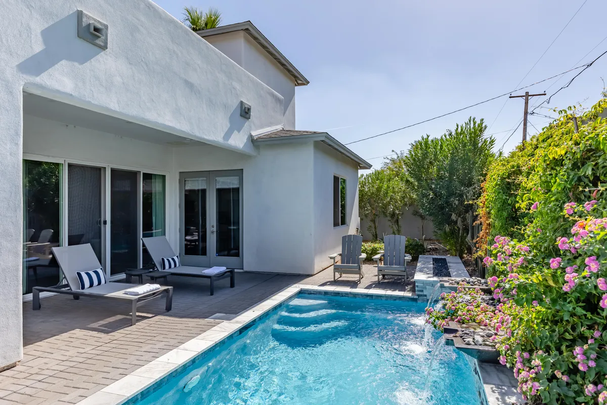 An outdoor scene featuring a sparkling swimming pool, modern lounge chairs, lush greenery, and vibrant flowering plants. The space is designed for swimming, sunbathing, outdoor dining, or hosting social gatherings, with an ambiance of relaxed luxury.