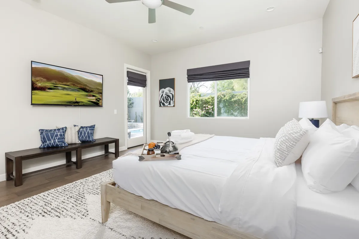 A bright and serene bedroom featuring a neatly made bed with white linens, a television mounted on the wall, decorative wall art, and a window letting in natural light. The room offers a sense of tranquility and comfort, ideal for relaxation.
