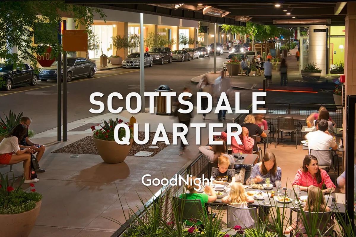 An illuminated outdoor dining and shopping area, featuring people enjoying al fresco meals under string lights, with storefronts and a lively street atmosphere. The scene emphasizes the inviting nightlife and social opportunities of the area.