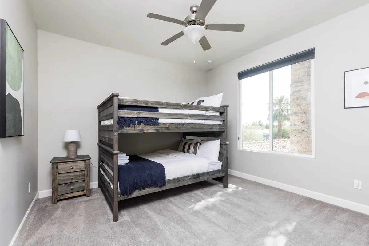 A playful and cozy bedroom with a wooden bunk bed, a nightstand with a lamp, and a window offering a pleasant view. The room is designed for both restful sleep and fun sleepovers, perfect for children or friends.